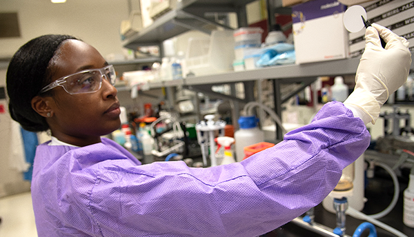 Alshae' Logan-Jackson wears a purple coverall and safety glasses in the lab as she uses tweezers to hold a small white disk. 
