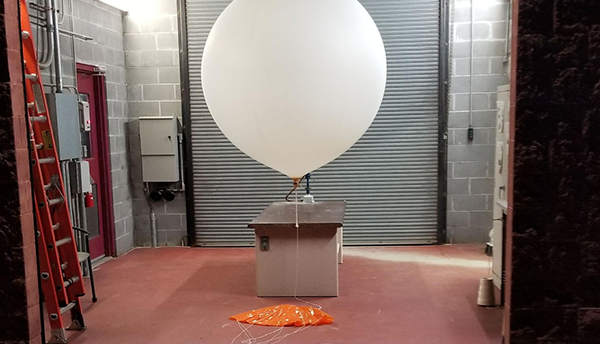 A large white balloon with a small box attached to it waits inside a cinderblock building with a rolltop door. 