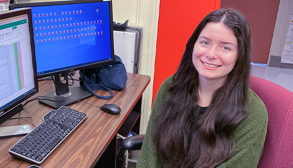Isabelle Rivera poses smiling at a computer with two monitors, one showing a spreadsheet and the other multiple file icons. 