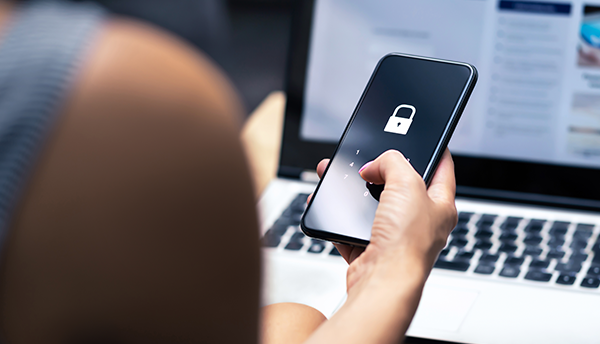 A person sitting at a laptop is holding a cellphone with a padlock showing on the screen.