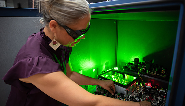Tara Fortier is wearing safety glasses as she reaches into a box of circuitry and other equipment, which emits a green glow. 