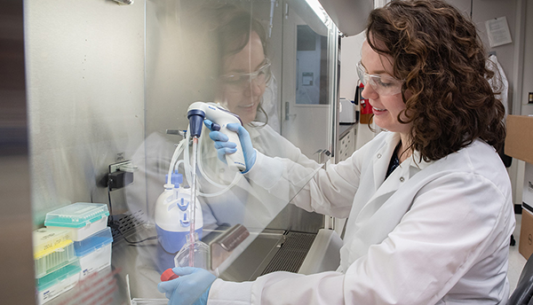 Jerilyn Izac sits at a lab bench with her hands inside a plastic shield, using a device to draw liquid from a plastic container into a tube. 