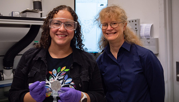 Diana Ortiz-Montalvo, wearing safety glasses and holding a small circular filter with tweezers, poses with Abigail Lindstrom in the lab.