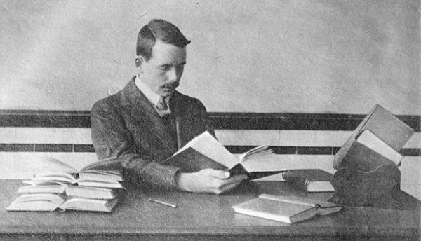 Historical photo shows Henry Moseley sitting at a desk reading a thick book, with other open books on the desk.