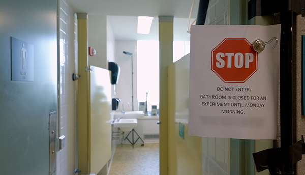 A paper sign is clipped to an open door to a public restroom. The sign reads, “Stop. Do Not Enter. Bathroom is closed for an experiment.”