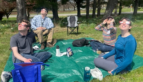 Four people sitting outside on a green tarp wear eclipse glasses as they peer up into the sky. 