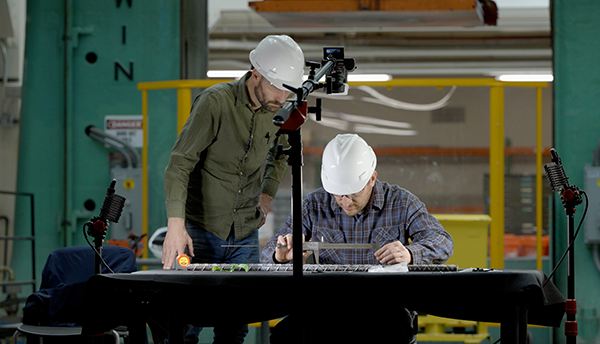 Two Champlain Towers researchers in hard hats lean over a rebar sample on a table in a large workspace. 