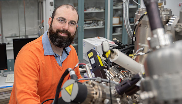 Trey Diulus, a bearded man in an orange sweater, smiles as he poses in the lab next to a complicated-looking scientific device.