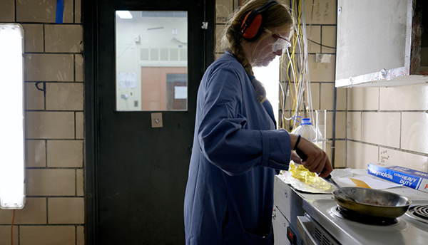 A researcher wearing safety glasses and hearing protection cooks bacon on a stovetop in a fire lab.