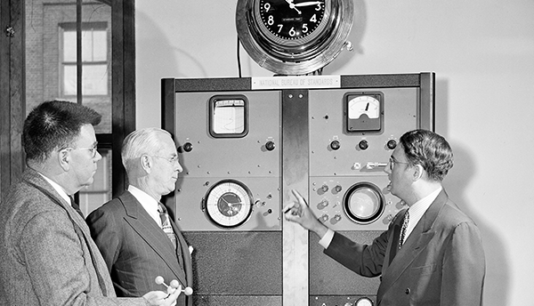 Historical photo shows three men standing next to a refrigerator-sized piece of machinery with an analog clock attached to the top. 