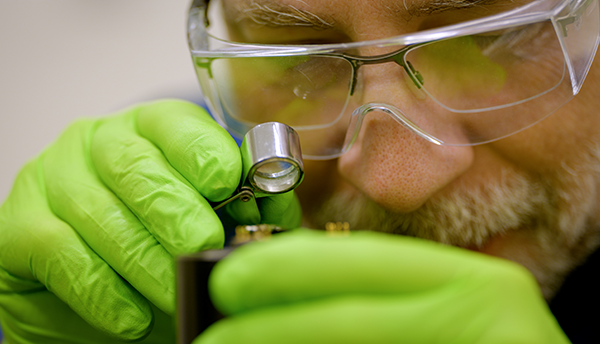 A researcher wearing green gloves and safety glasses over his regular glasses holds a small magnifying lens close to his face to look at a hard drive.