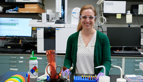 Briana Capistran wears safety glasses as she poses smiling in the lab with computers and other equipment in the background. 