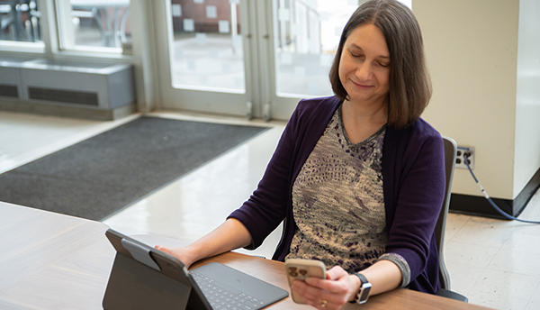 Julie Haney sits at a table in the NIST library with a laptop, looking at a smartphone in her hand. 
