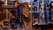 NIST-F4 researchers in safety glasses stand behind a table holding various electronic and laser devices that are part of the atomic clock. 