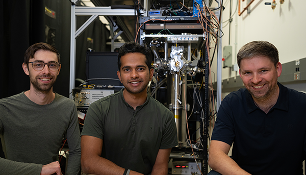 Three male researchers pose smiling in front of a large, complex random number generating device in a lab.