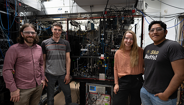 Four aluminum ion clock researchers pose standing around a large piece of scientific equipment covered with wiring. 