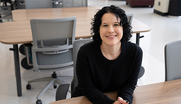 Kelly Sauerwein poses sitting at a table in the NIST library, leaning forward and smiling. 