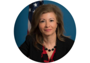Professional headshot of Azita Valinia, a woman with long brown hair, wearing a necklace, black blazer, and red top, smiling in front of a U.S. flag.