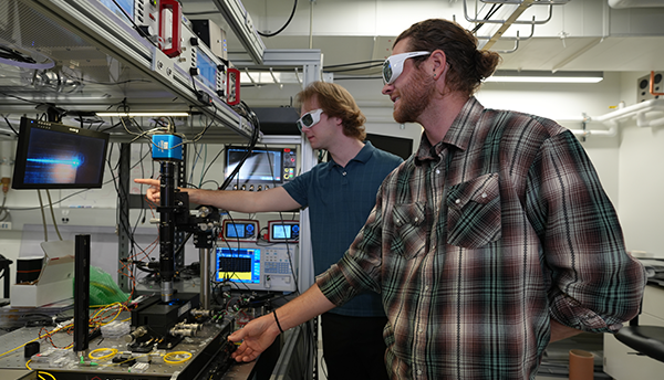 Two male researchers in protective eyewear stand in the lab, pointing at a screen hooked up to an any-wavelength laser. 