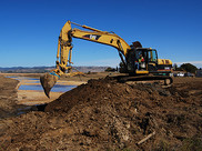 An excavator on a berm digs dirt to make a channel.