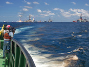 On the deck of a large boat, a videographer films the oiled waters of the Gulf of Mexico, with an oil platform in the background.