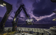 =A remotely operated vehicle is lifted onto a ship deck by a large mechanical arm.