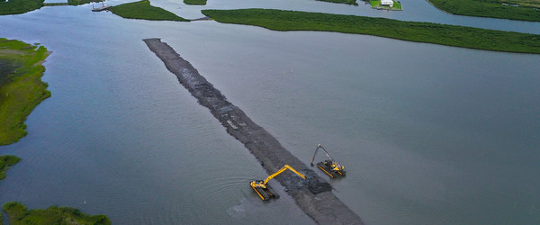 Aerial image of construction equipment moving dirt into a ridge surrounded by marsh and water.