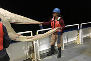 A young woman in work clothing and safety gear handles a fine-mesh plankton net at night on a research ship