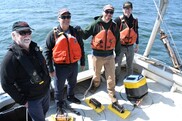 Four men wearing life jackets stand at the back of a boat. At their feet are a small yellow buoy and sensors attached along a wire.