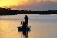 A man stands in a boat fishing at sunrise. The water reflects an orange hue and clouds. Credit: Florida Fish and Wildlife Conservation Commission