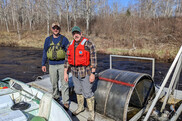 two men stand in a shallow skiff with wire trap in a marshy area