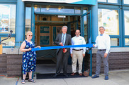 two people hole either end of a large ribbon at the doorway of a building while a third person cuts the ribbon with large scissors