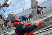 A person wearing a red life jacket and red rubber gloves stands on a ship pulling a rope.
