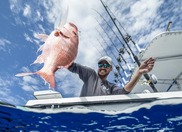 A man in sunglasses and a hat stands on a boat holding up a red fish. 