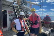 Two crew members stand side by side on a survey ship with life vests and hard hats on, both are smiling