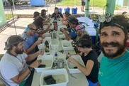 A group of researchers sits at a long outdoor table, each with a white plastic bin in front of them. Credit: ISER.