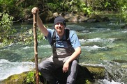 A person wearing waders sits next to a river holding a large branch.