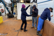 Two crew members hand off a Niskin bottle, as a third crew member holds a line with more attached over the side of the vessel. 