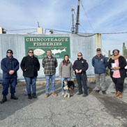 A dog and seven people of different ages, genders, and ethnicities stand in a parking lot.