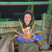 A field scientist in foul weather gear holds up a large crab aboard a commercial fishing vessel at night.