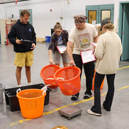 Four people with clipboards stand in a warehouse. Two are holding orange fish sorting baskets.