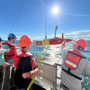 Men in hard hats pull orange long range AUV over side of ship at sea