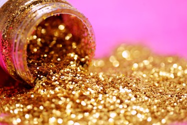 A gold jar of glitter is dumped out over a pink background.