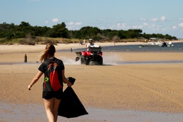 A person walking down a shoreline with a trash bag, other volunteers, boats, and a person on an all terrain vehicle are in the background.