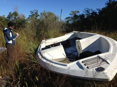 A researcher records data while standing near an abandoned vessel in tall grass.