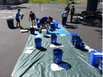 Labeled buckets are placed on a tarp on a blacktop and students are putting items into the buckets.