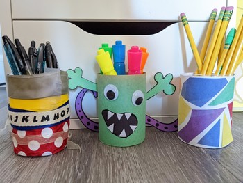 Three pencil cups with writing instruments. One is a green paper monster, one is decorated with ribbon, and one has a stained glass collage.