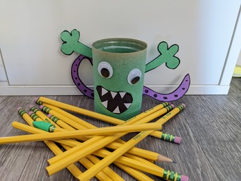 A cup decorated with a green paper monster surrounded by a mess of yellow pencils.