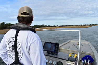 A person driving a boat equipped with a sonar screen. They are near a marsh edge.