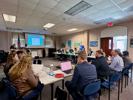 Advisory council members sit around a u-shaped table in front of a large screen during a meeting.